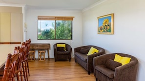 A living room with a wooden dining table, two sofas with yellow cushions, a window with a view of greenery, and a painting of sunflowers on the wall. - Spacious Unit overlooking Moreton Bay - Boyd St, W (WOORIM)