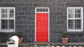 Un bâtiment avec une porte rouge, deux fenêtres à cadres blancs et deux plantes en pot.