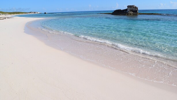 Une plage à l’eau turquoise et limpide, un rivage sablonneux et, au loin, un promontoire rocheux.