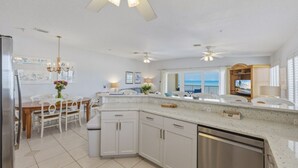 A modern kitchen with white cabinets, a stainless steel dishwasher, and a dining area with a wooden table and chairs. - Family-Friendly 3BR Beachfront Retreat | Indian Rocks | Sea Side #202 (Indian Rocks Beach)