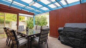 A covered patio with a glass table and chairs, a barbecue grill, and a wooden wall. - Family home located around the corner from Pea Soup (PORT FAIRY)