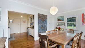A dining area with a wooden table and chairs, a kitchen in the background, and artwork on the walls. - A quaint artistic cottage for all seasons (PORT FAIRY)