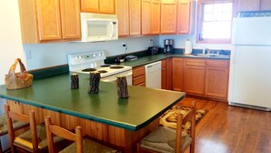 Una cocina con armarios de madera, una estufa blanca, un microondas y un lavavajillas. Hay cuatro sillas de madera con asientos de mimbre.