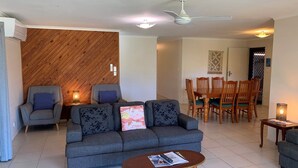 A living room with a grey sofa, two armchairs, a wooden accent wall, a dining area with wooden chairs, and a ceiling fan. - Lowset Pet Friendly Home (BONGAREE)