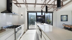 A modern kitchen with a gas stove, oven, and a large island with a sink. There is a dining area with a table and chairs visible through a glass door.