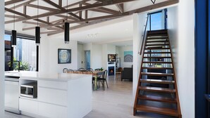 A modern kitchen with a central island, a dining area with chairs, a staircase, and a living room with a fireplace.