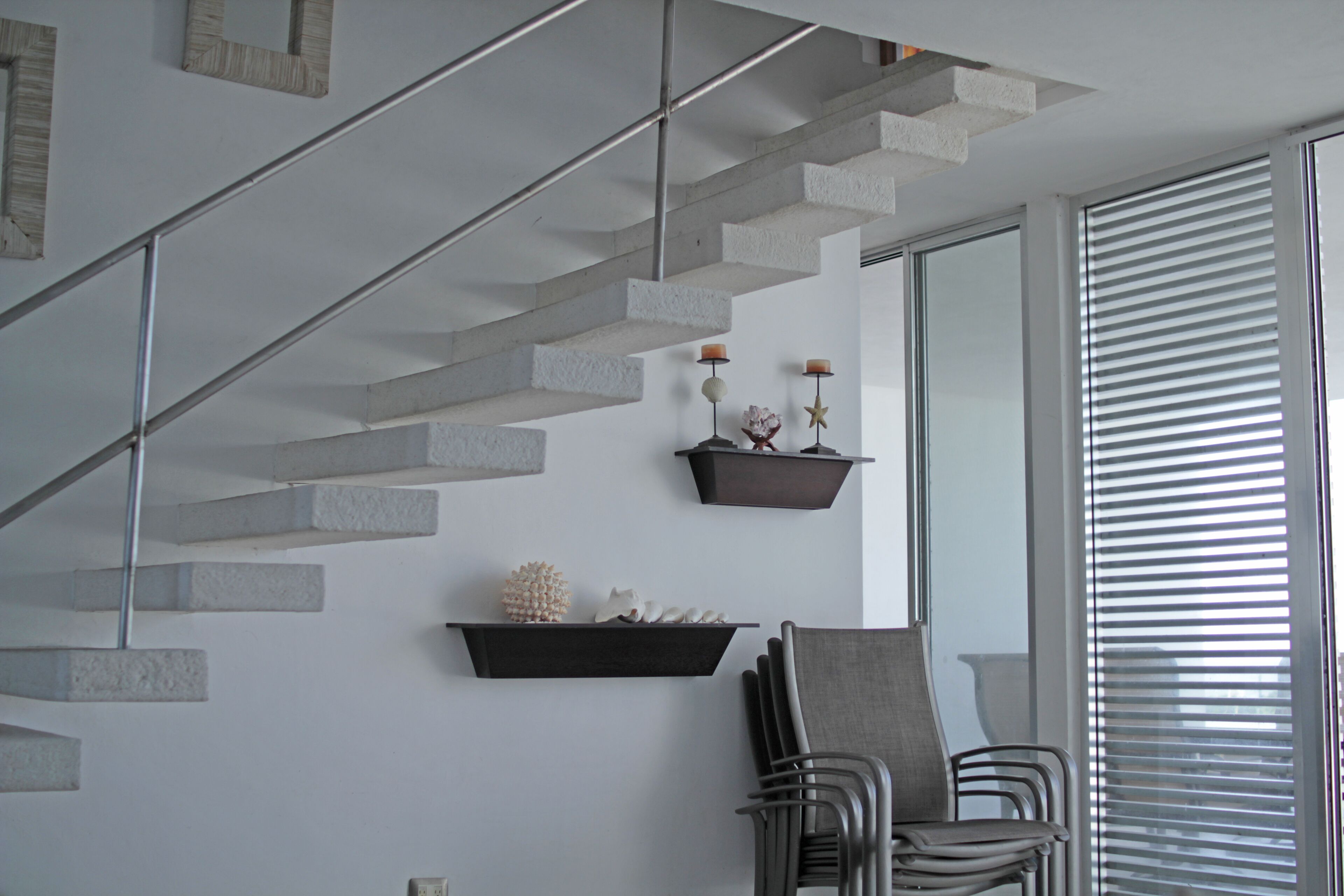 A modern interior with a concrete staircase, wall-mounted shelves with decorative items, and a seating area with chairs.