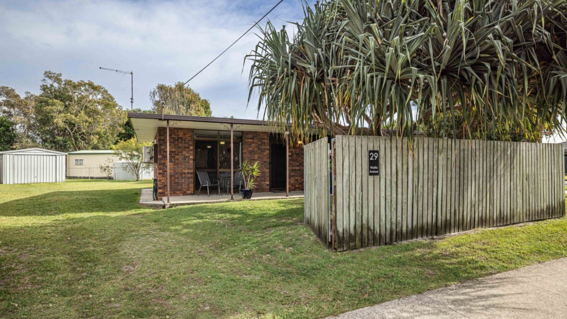 A single-story building with a patio, a fenced area with a sign '29 Wattle Avenue,' and a grassy yard.
