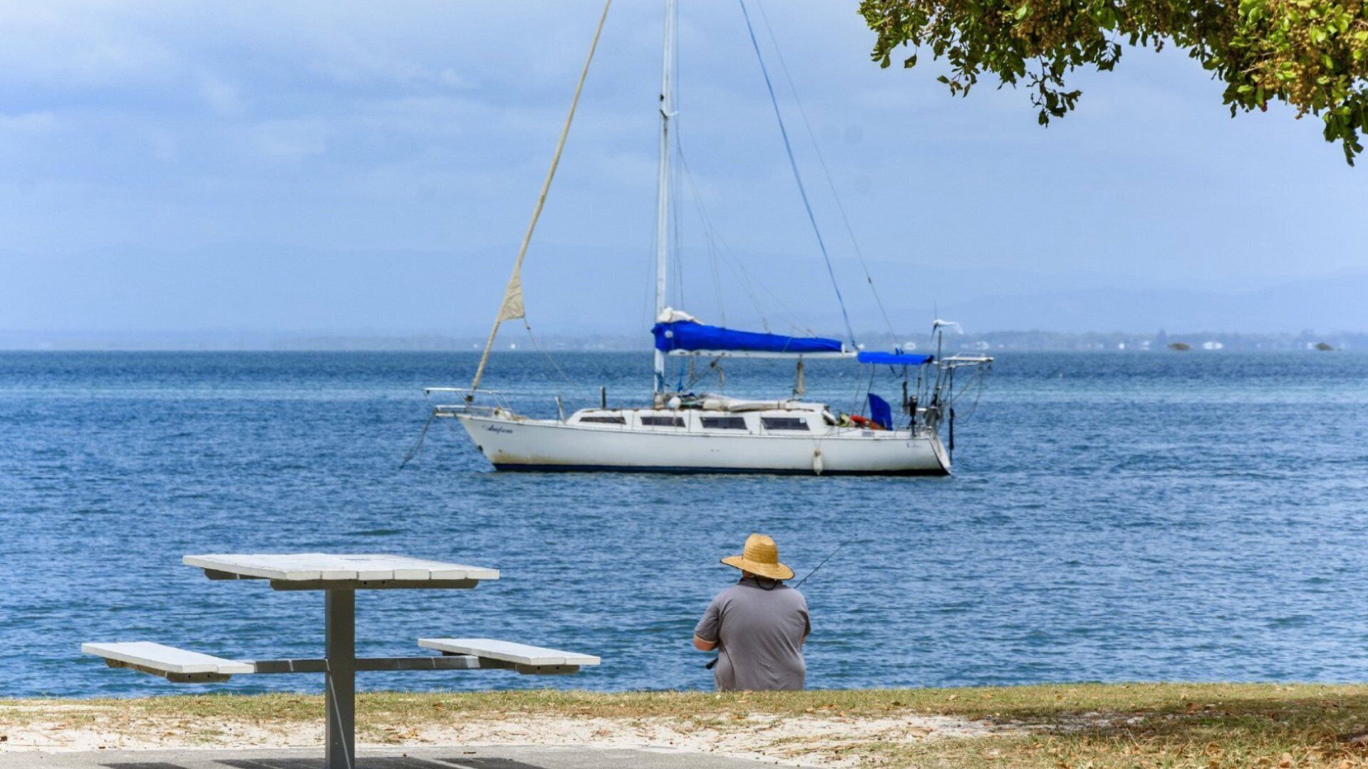 A person fishing by a calm sea with a sailboat in the background.
