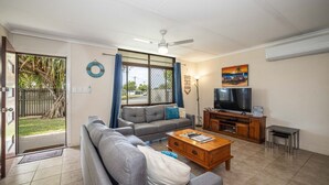 A living room with a sofa, coffee table, TV, and a view of the outside. - Bongaree Retreat With Room For The Boat (BONGAREE)