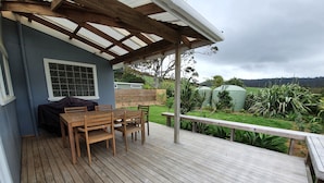 A wooden deck with a table and chairs, a covered seating area, and a view of a garden with plants and a green structure in the background.