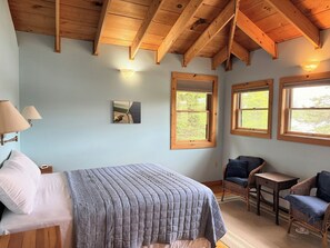 A bedroom with a bed, two windows, a chair, a table, and a lamp.