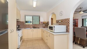 Een kleine keuken met een magnetron, fornuis en gootsteen. Er is een raam met uitzicht, en een klok aan de muur.