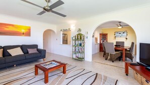 Een woonkamer met een bank, salontafel en televisie. Er is een eethoek met stoelen en een tafel. De kamer heeft een plafondventilator en een schilderij aan de muur.