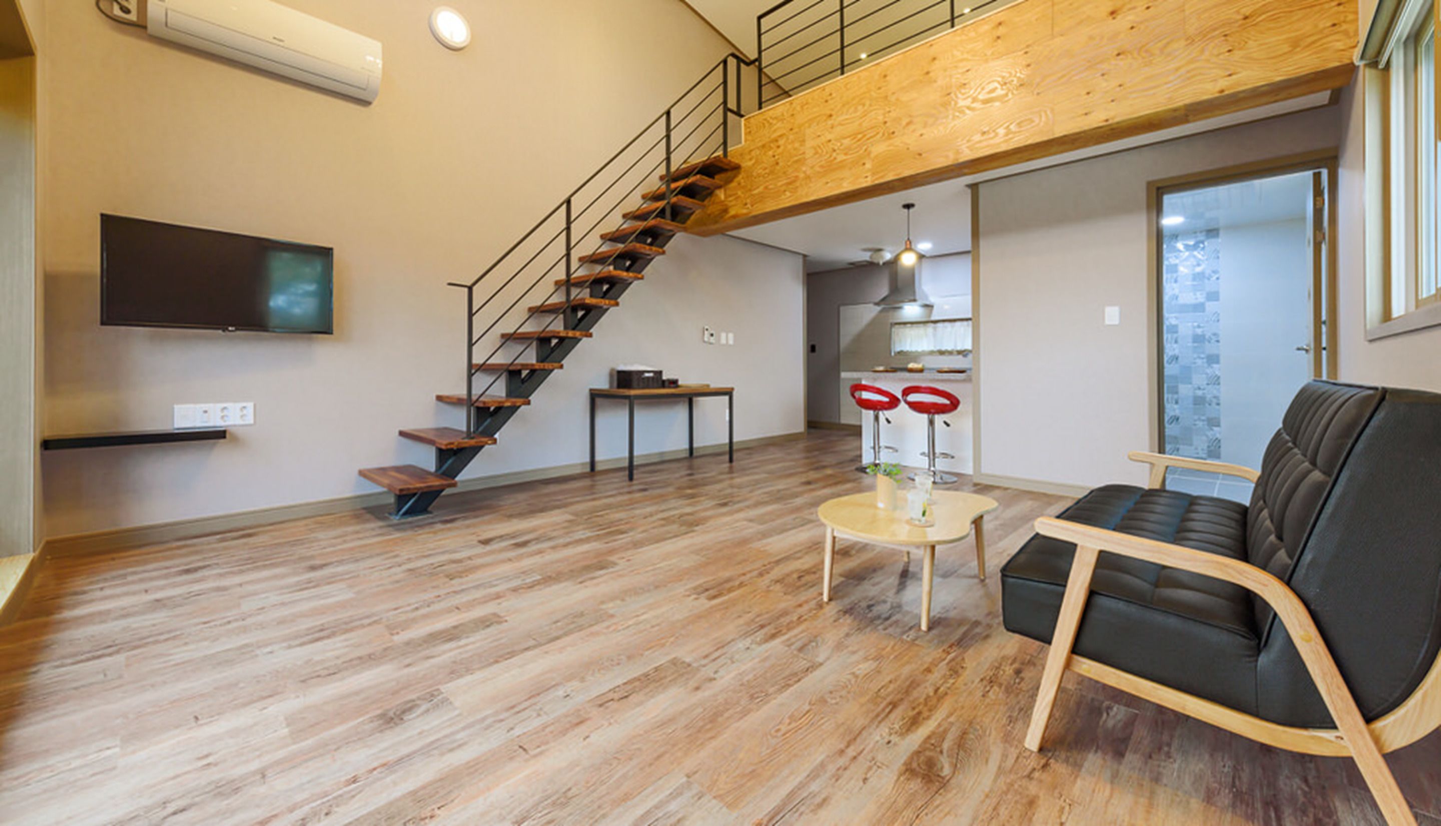 Un amplio espacio de sala con una escalera de madera, un televisor de pantalla plana, una mesa de comedor con dos taburetes rojos de cuero y un sofá de cuero negro.