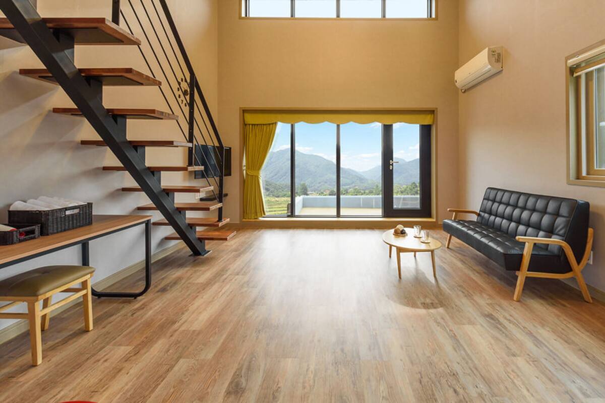Ein geräumiger Wohnbereich mit einer Holztreppe, einem schwarzen Ledersofa, einem kleinen runden Tisch und einem großen Fenster mit Blick auf die Berge.