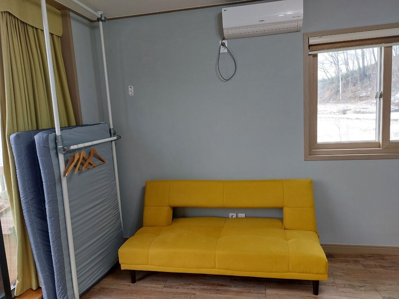 Et værelse med en gul sofa, en radiator, et vindue med udsigt til træer og en gardin.