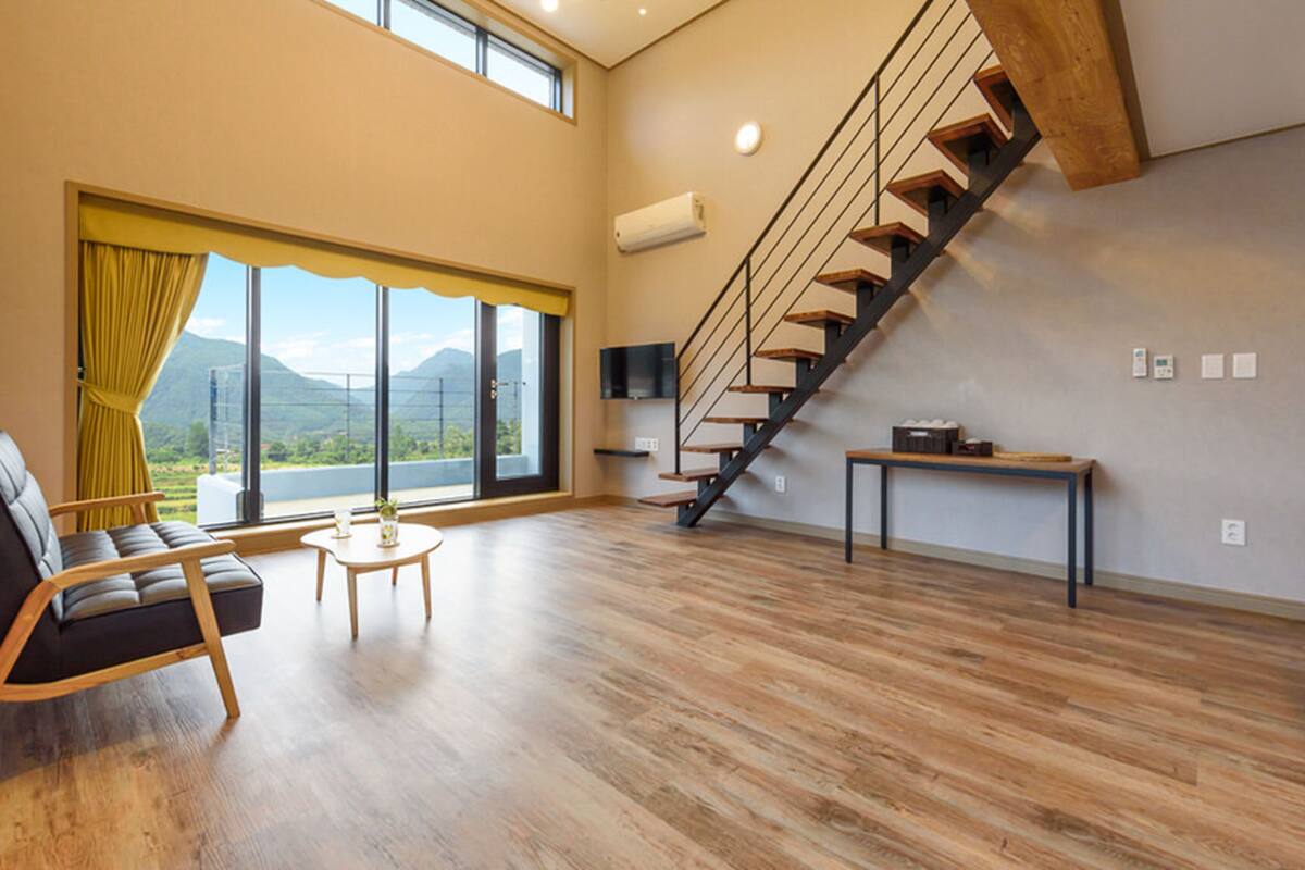Ein geräumiges Wohnzimmer mit Holzboden, einer Couch, einem kleinen Tisch, einer Treppe und einem großen Fenster mit Blick auf die Berge.