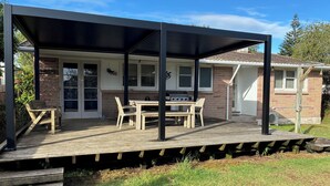A covered outdoor patio with a grill, wooden furniture, and a brick house in the background.
