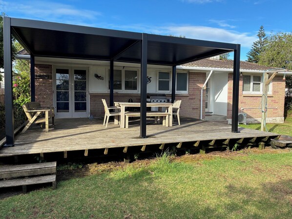 A covered outdoor patio with a grill, wooden furniture, and a brick house in the background. - Only 40 metres to the beach! (Ruakaka)