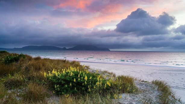 A beach with a view of the ocean and distant mountains under a colorful sky.
