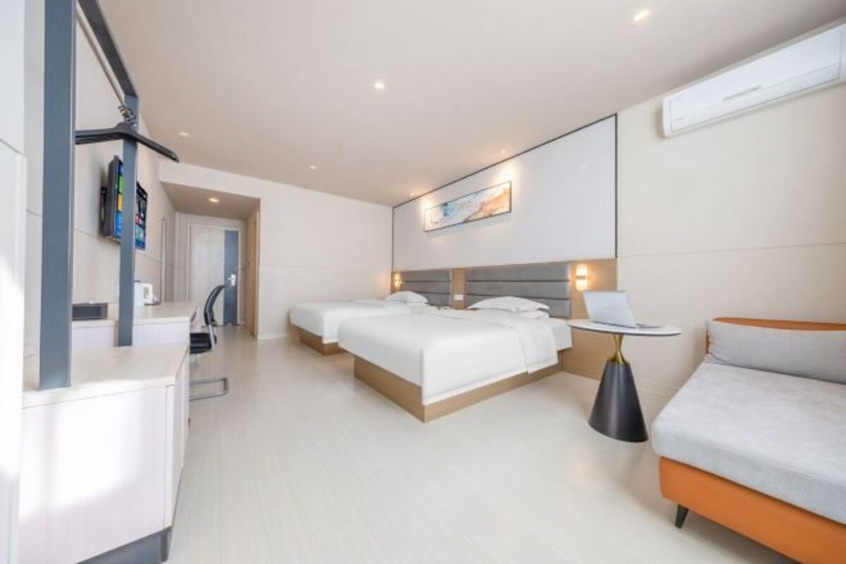 A modern hotel room with two beds, a sofa, a desk, and a small table with a lamp.