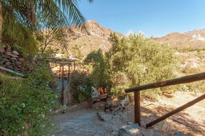 A rustic outdoor area with a stone wall, wooden pergola, and a small bridge leading to a shaded area.