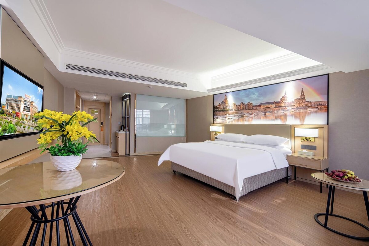 Habitación de hotel con cama, escritorio con silla, mesita con fruta, un cuadro de un paisaje urbano y ventana con vistas a edificios.