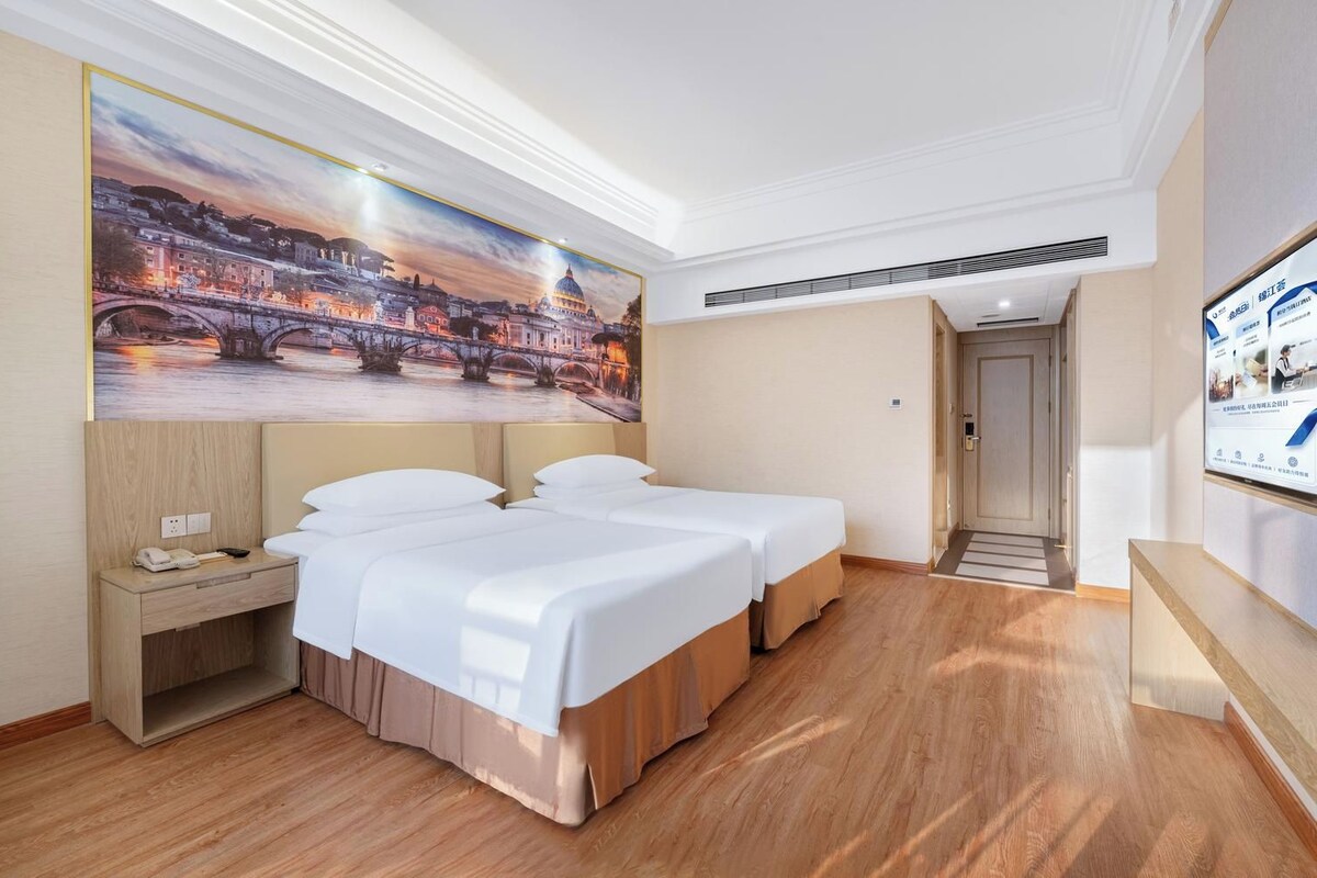 Habitación de hotel con dos camas, una mesita de noche, un cuadro de un paisaje urbano y piso de madera.