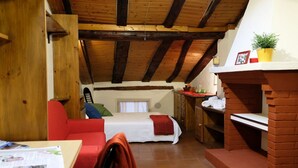 A cozy room with a bed, a red sofa, a small table with a newspaper, a brick fireplace, and a window with curtains.