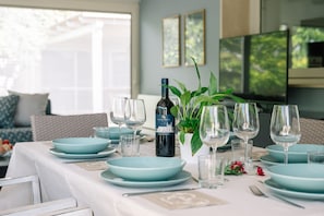 Una mesa de comedor dispuesta con platos azules, copas de vino y una botella de vino, con una planta y un televisor al fondo.