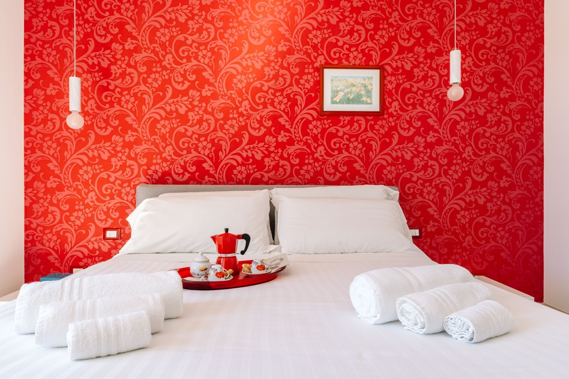 A hotel room with a red patterned wallpaper, a bed with white linens, a tray with a teapot and cups, and rolled towels.