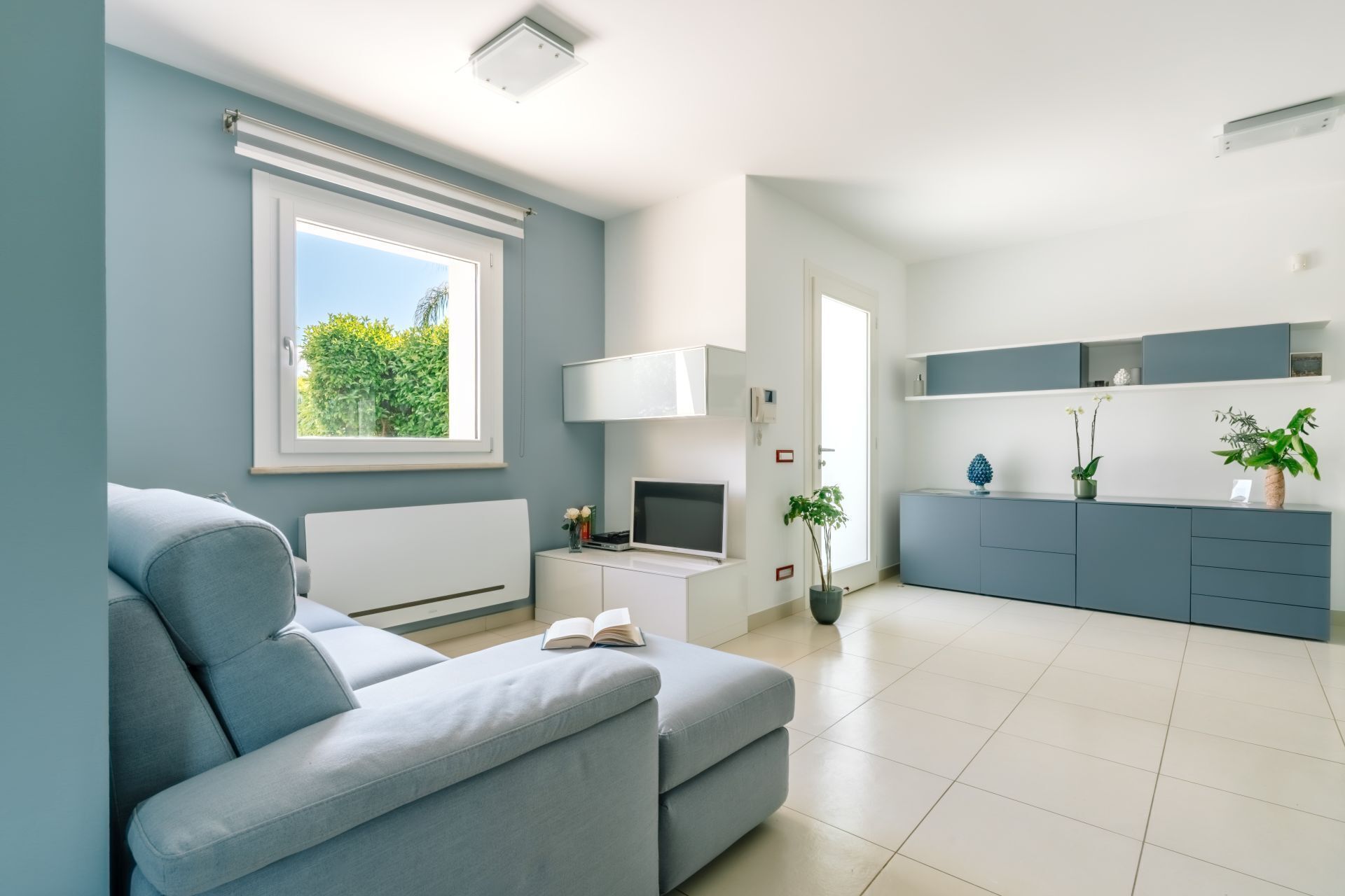 A modern living room with a sofa, a TV, and a window with a view of greenery.