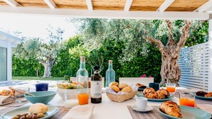 Un espacio de comedor al aire libre con una mesa dispuesta para el desayuno, incluyendo pan, bollería y bebidas.