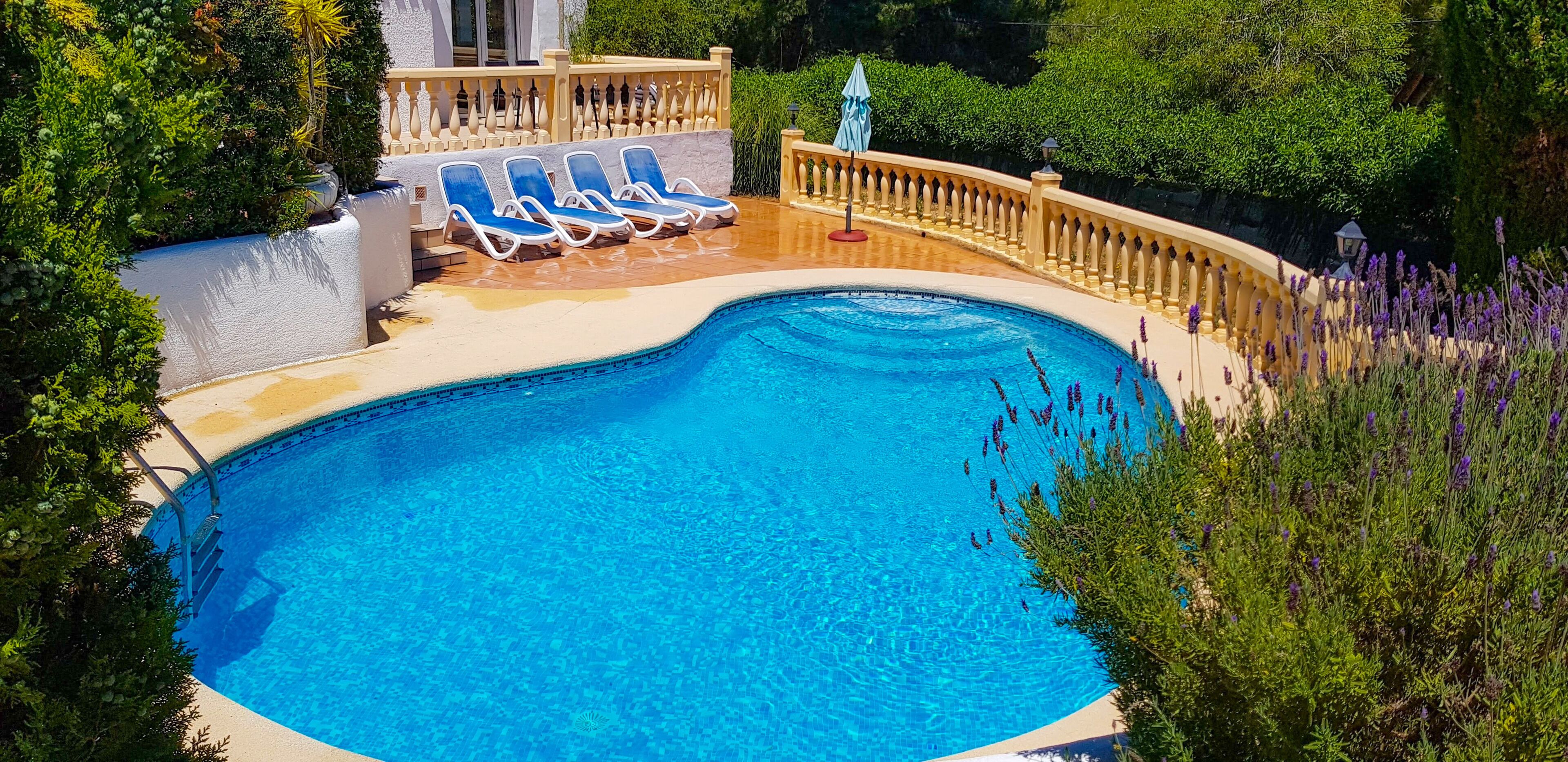 Una piscina con agua azul, rodeada por una terraza color beige y una pared blanca. Hay sillas reclinables azules y un paraguas rojo.