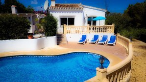 A swimming pool with clear blue water, surrounded by a beige deck and a white wall. There are five blue lounge chairs aligned along the deck.