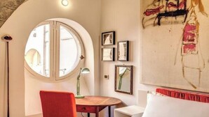 A room with a round window, a desk, a chair, and a bed. There are framed pictures on the wall and a large abstract painting.