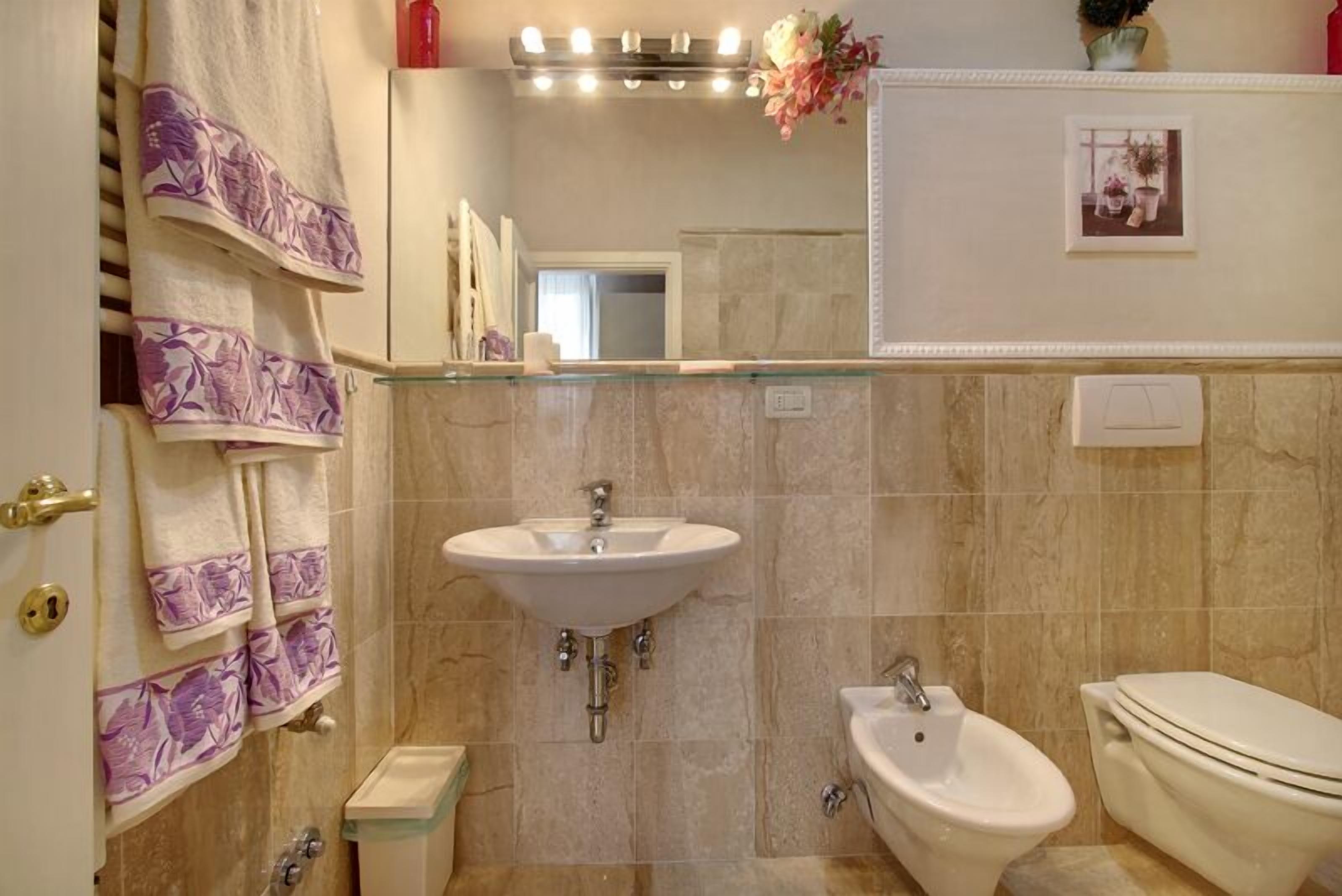 Een badkamer met een wastafel, bidet en toilet. Handdoeken hangen aan een rek, en boven de wastafel is een spiegel.