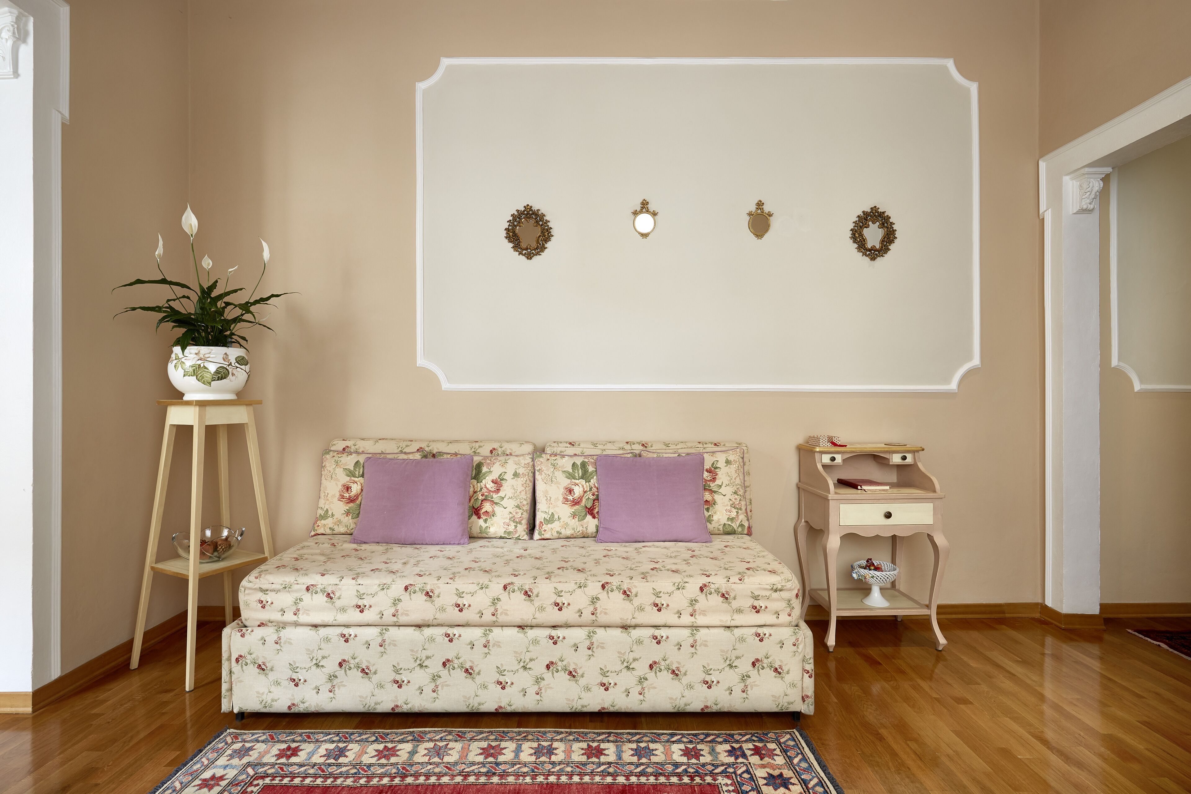 A bedroom with a floral sofa, a small wooden side table, a decorative wall piece, and a patterned rug.