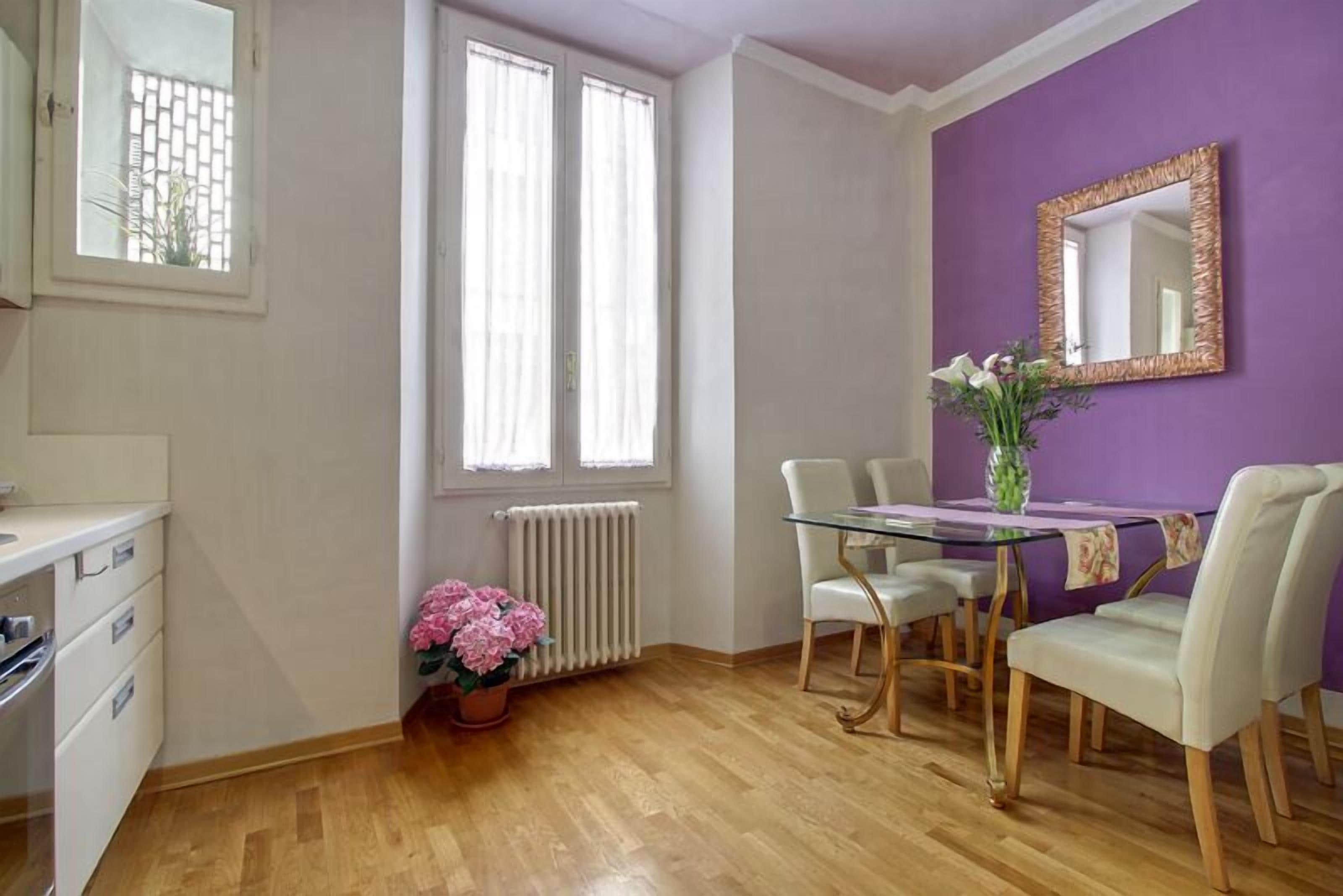 A dining area with a glass table, white chairs, and a purple wall.