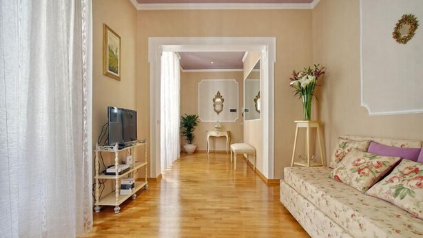 A living room with a wooden floor, a sofa with floral upholstery, a television on a stand, a small table with a plant, and a mirror on the wall. - The elegance in the heart of Florence (Firenze)