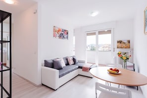 A modern living room with a white sofa, a dining table with chairs, and a balcony with a view.