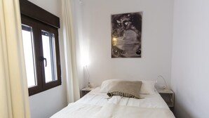 Een slaapkamer met een bed, nachtkastje, gordijnen voor het raam en een kunstwerk aan de muur.