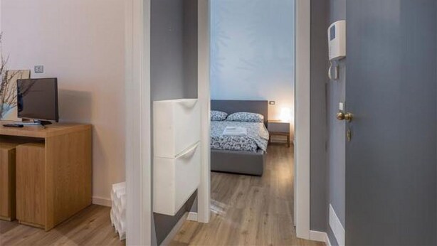 Ein Flur, der zu einem Schlafzimmer führt, mit einem Bett, einem Schreibtisch mit Computer und einer Tür mit Lichtschalter.