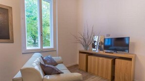 Ein Wohnzimmer mit einer Couch, einem Fernseher auf einem Holzboden und einem Fenster mit Blick auf Bäume.