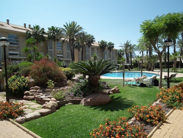 Pátio de hotel com piscina, palmeiras e um jardim bem cuidado com diversas plantas e flores.