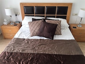 Uma cama bem arrumada com um edredom marrom, duas mesas de cabeceira com luminárias e uma cabeceira de madeira.