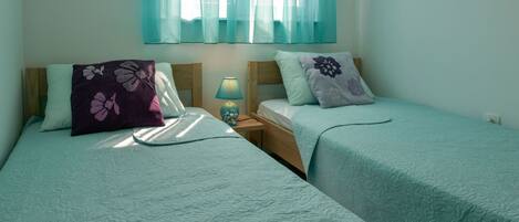A twin room with two beds, each with a floral pillow, a bedside table with a lamp, and sheer curtains.