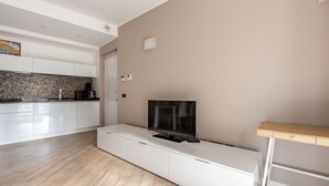 Una sala de estar moderna con un televisor de pantalla plana sobre un mueble blanco, una cocina con armarios blancos y un salpicadero de azulejos, y un suelo de madera.