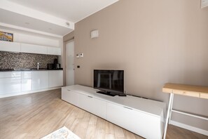 A modern living room with a flat-screen TV on a white entertainment unit, a kitchen with white cabinets and a tiled backsplash, and a wooden floor.